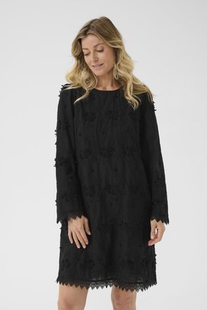 Femeie cu părul blond ondulat, purtând o rochie neagră cu mâneci lungi, cu modele florale texturate și dantelă la tiv și la mâneci.