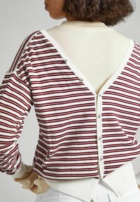 Pull rayé crème et bordeaux, avec un dos en V, une fermeture à boutons, et un tissu texturé avec des accents volantés sur les manches.