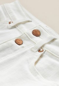 Weißer Denim-Stoff mit Kupferknöpfen, sichtbaren Nähten und einer kleinen Stoffschlaufe am Bund, die ein sauberes, minimalistisches Design präsentiert.