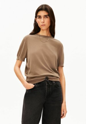 Femme aux longs cheveux foncés portant une chemise marron à manches courtes et un pantalon noir taille haute, debout avec une main dans la poche sur fond blanc.
