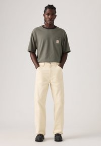 Ριγέ πουκάμισο με κοντά μανίκια σε ελιά και μαύρο χρώμα με μια μικρή τσέπη, συνδυασμένο με ανοιχτόμπεζ παντελόνι με ίσια γραμμή και μαύρα αθλητικά παπούτσια.