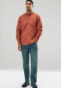 Rust-colored button-up shirt with a chest pocket, paired with light blue denim jeans and black slip-on shoes, standing against a neutral background.