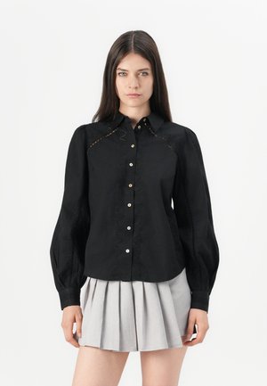 Black button-up shirt with long puff sleeves, featuring embroidered detailing along the seams and pearlescent buttons, paired with a grey pleated skirt.
