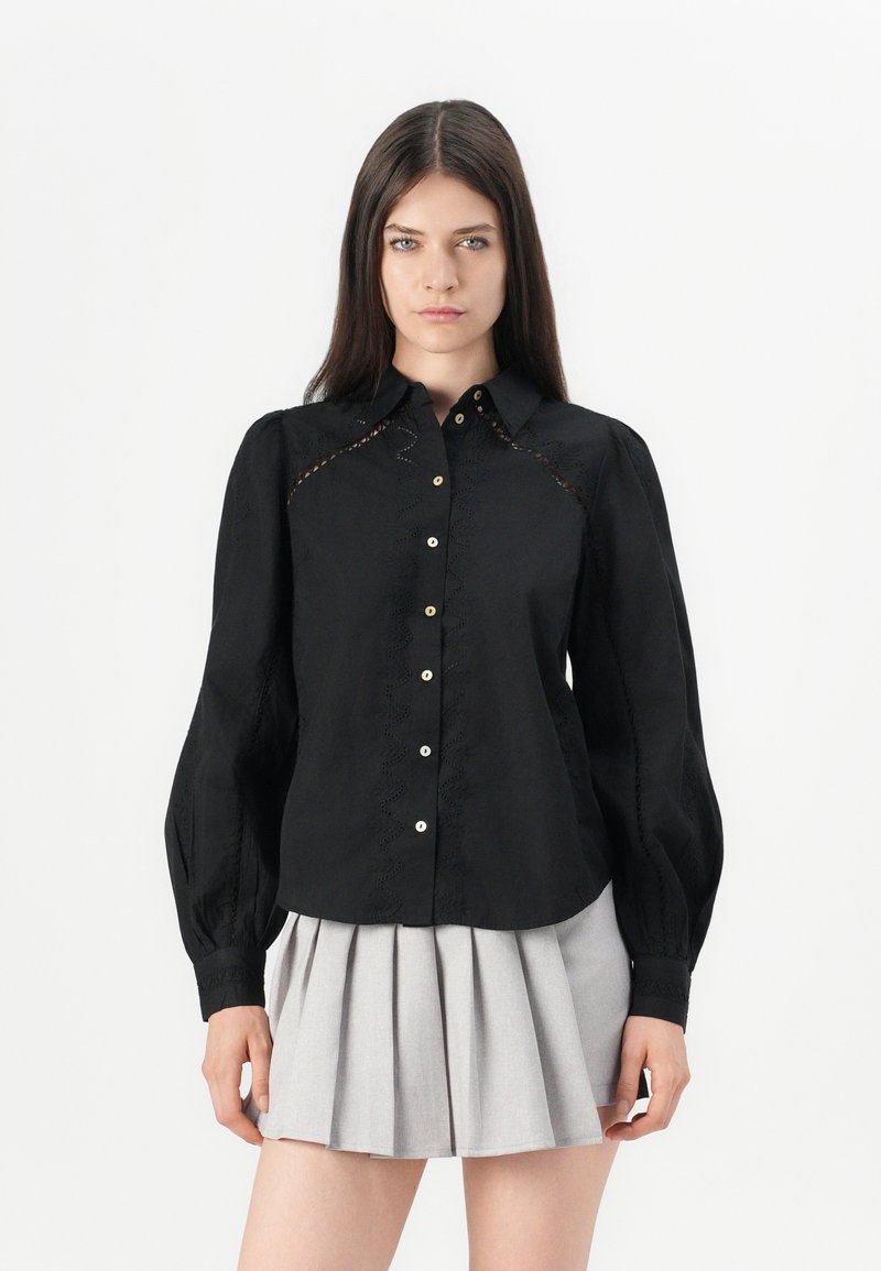 Svart skjorte med lange puffermer, med broderte detaljer langs sømmene og perleaktige knapper, kombinert med et grått plisserte skjørt.