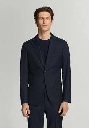 Uomo con capelli corti e scuri, che indossa una giacca da abito blu scuro aderente e pantaloni abbinati, in piedi davanti a uno sfondo chiaro e uniforme.