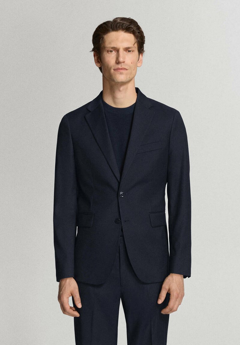 Homme aux cheveux courts et foncés, portant une veste de costume bleu marine cintrée et un pantalon assorti, debout devant un fond clair uni.