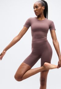 Καφέ κορμάκι με κοντά μανίκια και εφαρμοστή σχεδίαση, κατασκευασμένο από ελαστικό ύφασμα, συνδυασμένο με σορτς ποδηλασίας που φτάνουν μέχρι το μέσο των μηρών. Λείο υφή.