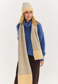 Modrý pletený rolák, šedý šátek se žlutým akcentem a béžová pletená čepice. Měkká textura, teplý design, vrstvený vzhled.