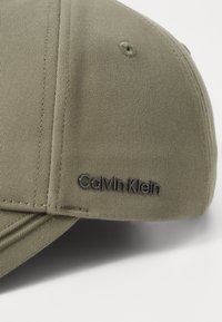 Olivengrønn baseballcap med bøyd skjerm, med en sydd tekstur og en svart "Calvin Klein"-logo på forsiden.