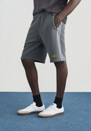 Sivé teplákové šortky s bielymi bočnými pruhmi a žltým nápisom "ORIGINALS"; doplnené bielymi teniskami s svetlosivými detailmi a čiernymi ponožkami.