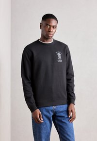 Czarny sweatshirt wykonany z bawełny, z ściągaczami przy mankietach i dolnym brzegiem. Na piersi znajduje się biała marka. W połączeniu z niebieskimi dżinsami.