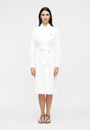 Γυναίκα με μακριά σκούρα μαλλιά που στέκεται, φοράει λευκό φόρεμα με μακριά μανίκια και ζώνη στη μέση, συνδυασμένο με μαύρα επίπεδα σανδάλια με λουράκια.