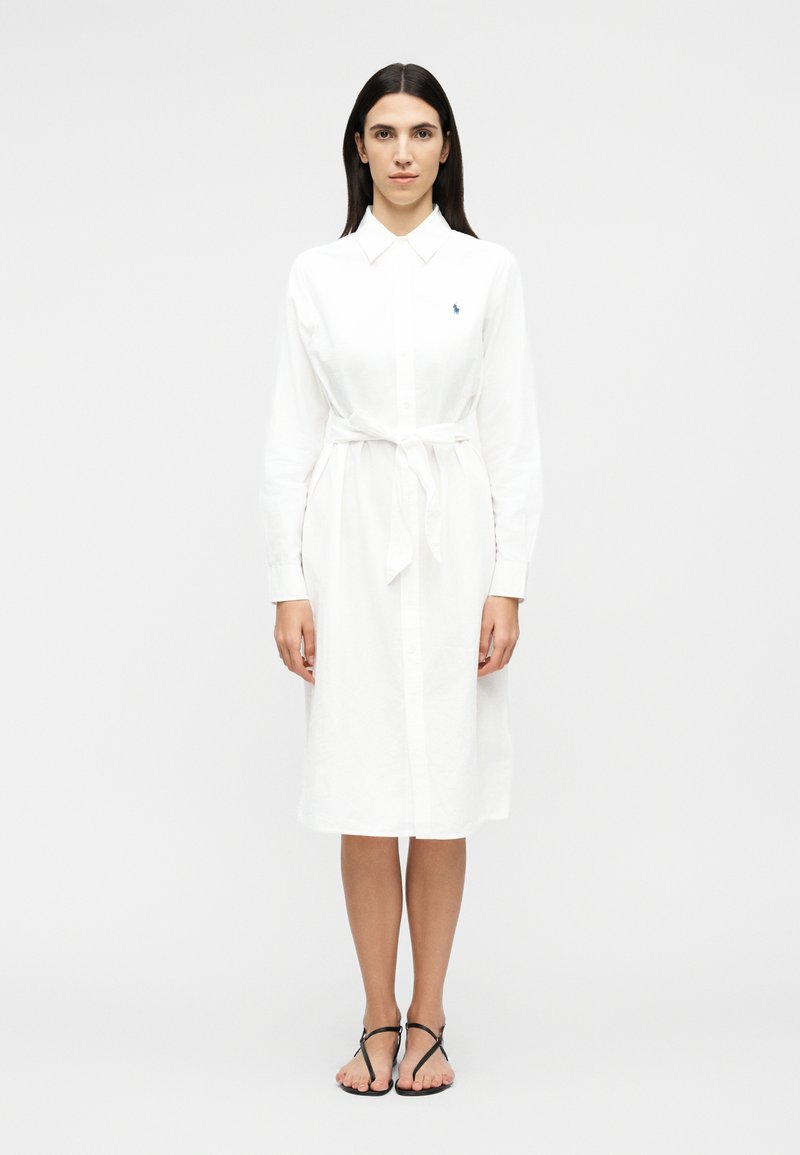 Femeie cu păr lung și întunecat care stă în picioare, purtând o rochie-tip cămașă albă cu mâneci lungi și un cordon din talie, alături de sandale plate negre cu barete.