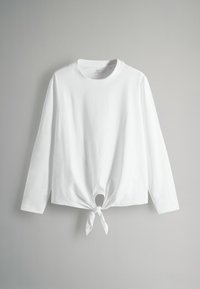 Camicia bianca a maniche lunghe in cotone, con scollatura rotonda e un dettaglio annodato in vita con una piccola apertura.