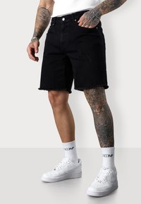 Black denim shorts with frayed hem and distressed details, worn with white sneakers and white socks. Tattooed leg visible on the right.