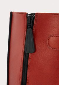 Gros plan sur une tirette de fermeture éclair noire sur une botte en cuir rouge avec des coutures visibles et une texture lisse.