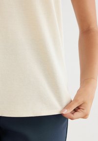 Hellbeiges ärmelloses Oberteil aus weichem Stoff, mit glatter Textur und leichtem Stretch, mit geradem Saum und seitlichen Nahtdetails.