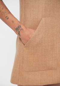 Tan ullblandat plagg med en stor stuckficka. Texturen verkar mjuk och något strukturerad, med ett subtilt mönster.