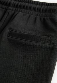 Black textured fabric joggers feature an elastic waistband and a small rear pocket with a rectangular flap.