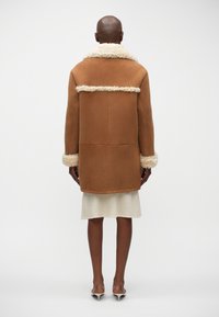 Chaqueta de piel de oveja marrón con un suave cuello y puños crema, que presenta una forma cuadrada y un diseño panelado en la parte posterior.