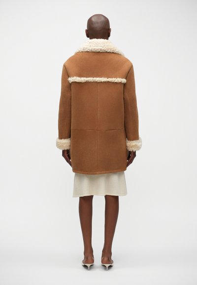 Giacca in shearling marrone con un morbido colletto e polsini crema, caratterizzata da una forma squadrata e un design a pannelli sul retro.