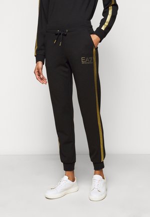 Zwarte joggers met gouden zijstrepen en EA7 Emporio Armani-logo, gedragen met witte sneakers, één hand in de zak.