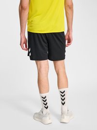 Hombre con camisa amarilla, pantalones cortos negros con rayas blancas en forma de chevrón, calcetines blancos con patrones negros en forma de chevrón y zapatillas grises, de pie de espaldas.