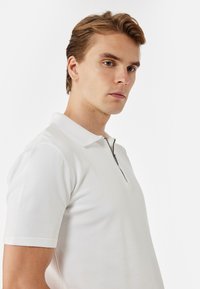 Ung man med ljusbrunt hår som bär en vit kortärmad polo med dragkedja fram, som tittar något åt sidan mot en vit bakgrund.
