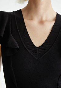 Haut en maille noire avec un col en V et des manches courtes à volants. Doté d'un tissu texturé et d'une coupe ajustée qui met en valeur des lignes épurées.