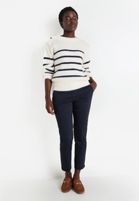 Pull crème avec des rayures marine, texture tricotée, détails de boutons sur l'épaule ; associé à un pantalon marine et des mocassins en daim marron.