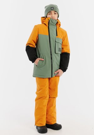 Orange- und grüne Jacke mit schwarzen Akzenten, ausgestattet mit einer Fronttasche und Reißverschluss, kombiniert mit orangefarbenen Hosen und schwarzen Stiefeln.