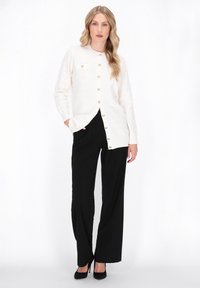 Cardigan bianco lavorato a maglia con scollatura rotonda, bottoni frontali e una leggera texture a righe; abbinato a pantaloni neri a gamba larga e tacchi.