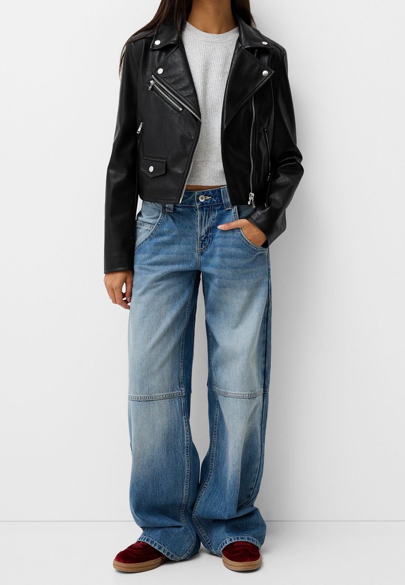 Černá kožená motorkářská bunda se stříbrnými zipy, šedý žebrovaný svetr, světle modré široké džíny a červené semišové boty.