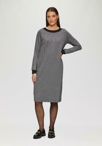 Harmaa ruudullinen mekko, jossa on pitkät hihat, pyöristetty musta kaulus ja mustat hihansuut. Yhdistetty mustien lakatunahan kenkien ja sukkahousujen kanssa.