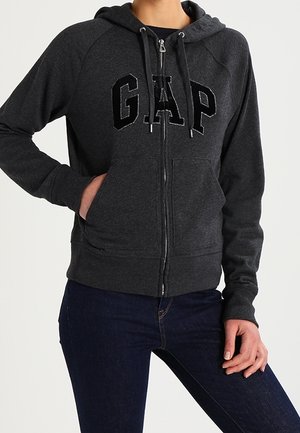 Person iført en mørkegrå hættetrøje med lynlås og GAP-logo foran samt mørkeblå jeans, med den ene hånd i lommen, foran en ensfarvet baggrund.