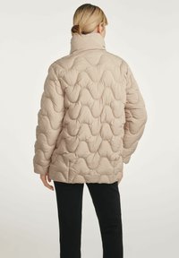 Beżowa kurtka puchowa z falistym pikowanym wzorem, wysokim kołnierzem i długimi rękawami, noszona na czarnych spodniach, prezentująca gładką teksturę i wyściełany design.