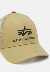 Haki kravas cepure ar noapaļotu vizu, sietu malām un melnu "ALPHA INDUSTRIES" logotipu, kurā ir stilizēts burts "A" un svītras.
