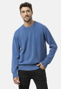 Blauwe gebreide trui met een ronde hals, ribgebreide mouwen en zoom. Gemaakt van een zachte stof en met een subtiele structuur. Draagbaar boven zwarte broek.