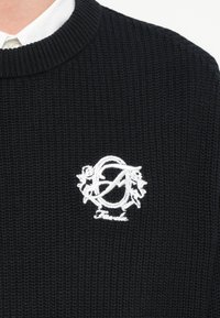 Črni pleteni pulover z okroglim izrezom in belim vezenim logotipom z rožnatimi poudarki na predelu prsnega koša.