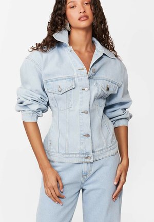 Kobieta o kręconych włosach, ubrana w jasnoniebieską kurtkę dżinsową i pasujące do niej jeansy, stoi z jedną ręką opartą blisko biodra.