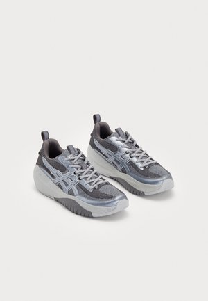 Paire de baskets grises et argentées avec semelles texturées et design à lacets sur un fond clair uni.