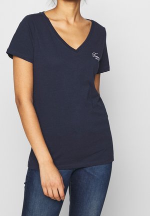 Donna che indossa una maglietta a maniche corte blu navy con scollo a V e jeans blu, in piedi con una mano rilassata al suo fianco.