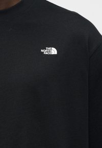 Κοντινό πλάνο μαύρου υφάσματος με μικρό λευκό κεντημένο λογότυπο «The North Face» στο στήθος ενός ρούχου με στρογγυλή λαιμόκοψη.