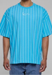 Mies, który ma na sobie luźną, jasnoniebieską koszulkę z cienkimi białymi pionowymi paskami i małym białym logo na piersi, w połączeniu z niebieskimi dżinsami.