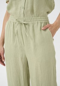 Pantalons en lin vert clair avec taille élastique et cordon de serrage. Comprend des poches latérales et un tissu texturé. Fermeture à bouton à la taille.