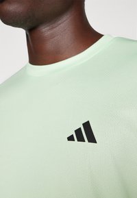 Hellgrünes Sport-T-Shirt aus strukturiertem Stoff, mit einem schwarzen dreieckigen Logo auf der linken Seite der Brust.