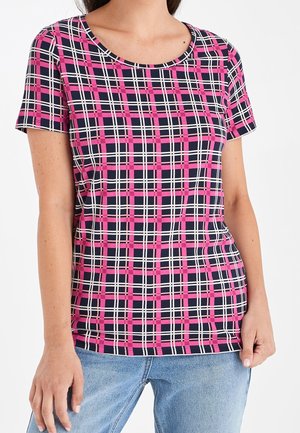 Femme portant un T-shirt à manches courtes avec un motif à carreaux rose, bleu marine et blanc, et un jean bleu clair sur fond blanc uni.