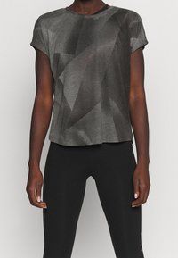 Persona con una camiseta de manga corta de rayas grises y negras, combinada con leggings negros sólidos, de pie frente a un fondo sencillo.