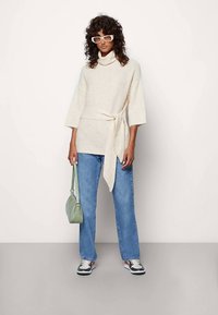 Cream knit sweater with a cowl neck and belted waist, paired with light blue straight-leg jeans and a small green handbag.