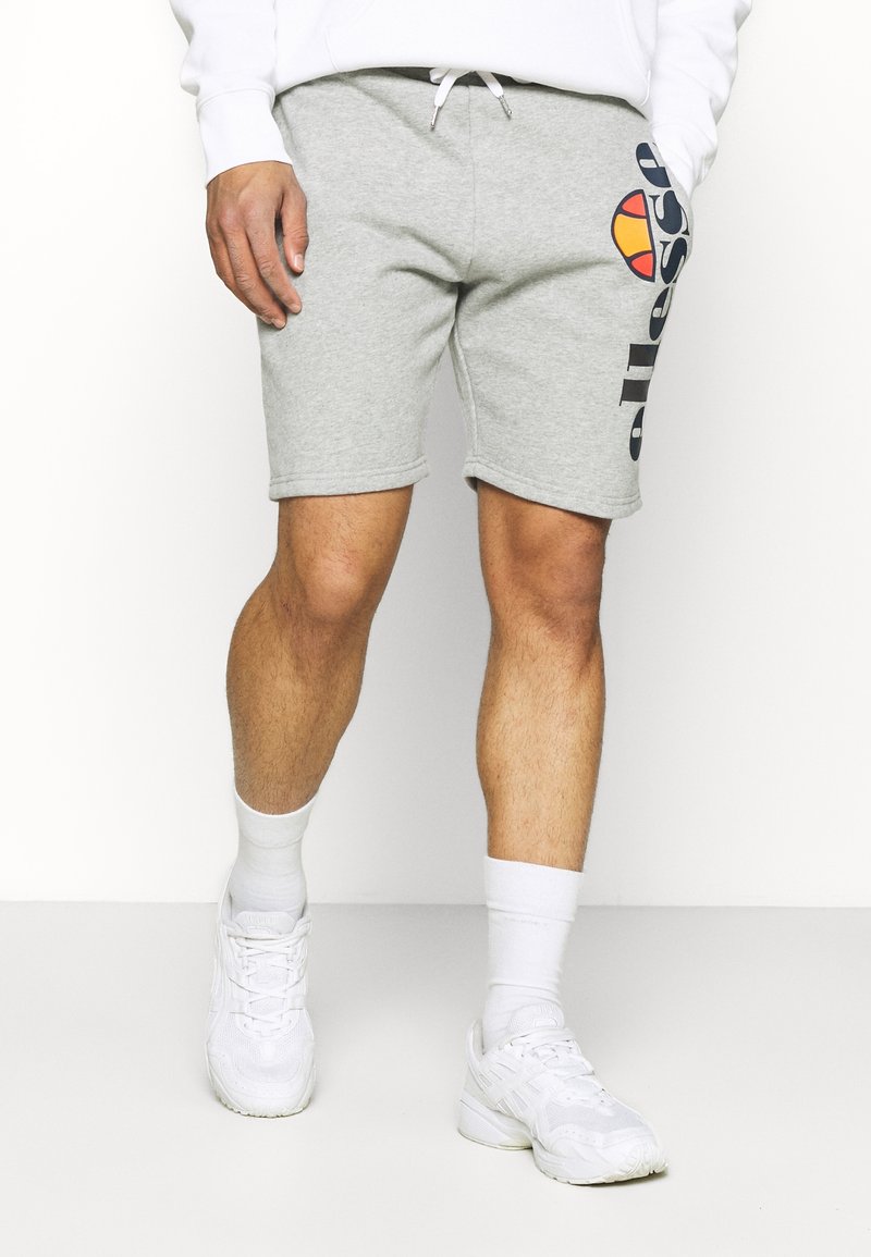 Homme portant un short de survêtement gris clair avec un logo Ellesse coloré, des chaussettes blanches et des baskets blanches, debout devant un fond uni.